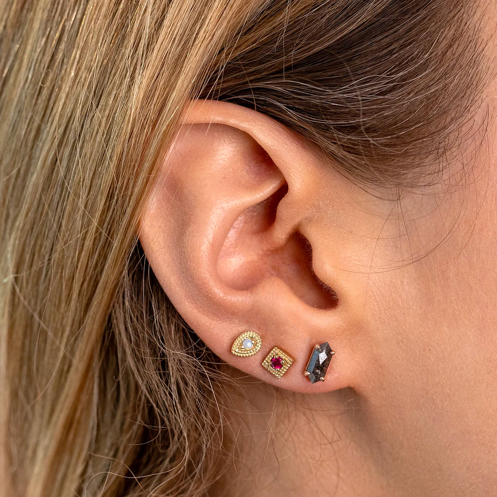 Close-up of three stud earrings by Emily Gill Jewelry, showcased on an ear. Features a pear-shaped opal with a diamond halo, a square ruby with a gold milgrain frame, and a geometric salt and pepper diamond in 14k yellow gold. Fine jewelry designed by Emily Gill, available at Ruby Mardi in Montreal, Canada.
