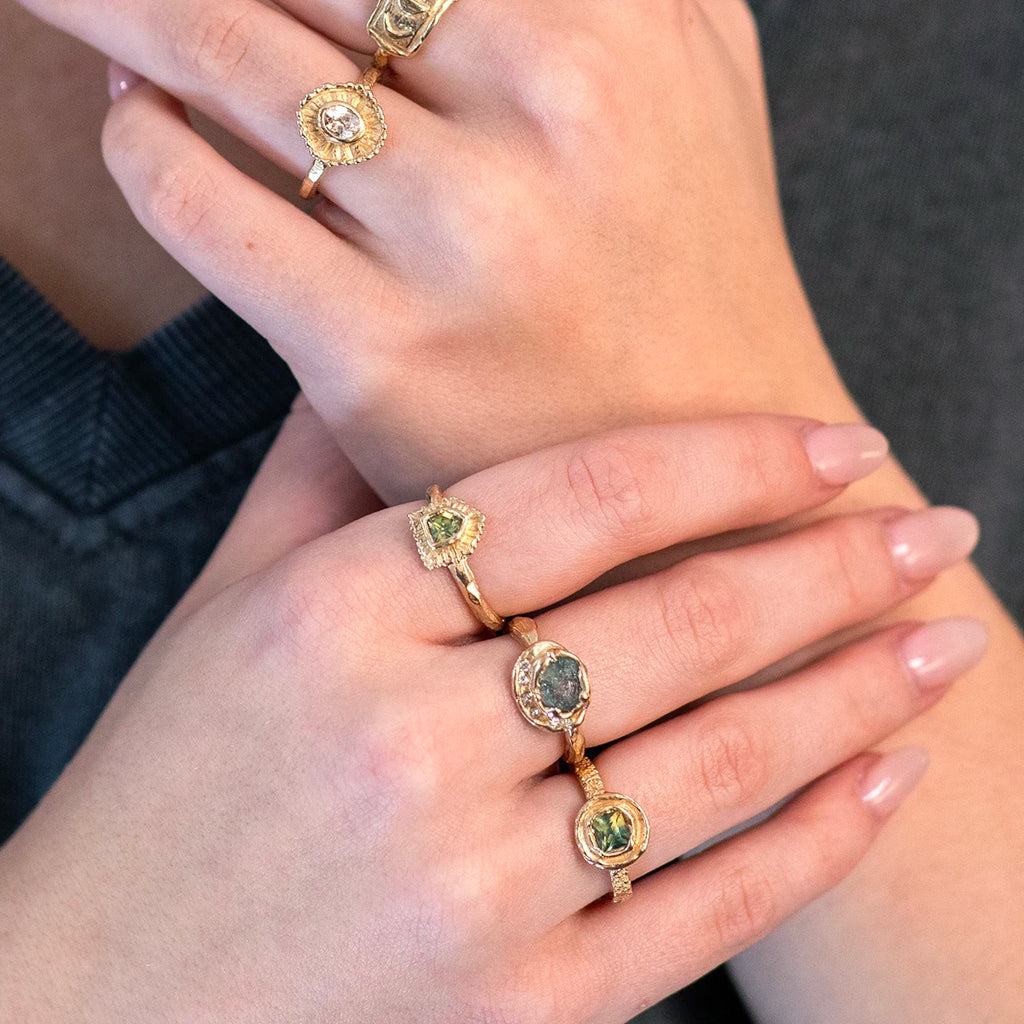 Close-up of four exclusive Emma Glover rings for Ruby Mardi, worn on both hands. One hand features a yellow gold ring with a white sapphire framed by a textured gold halo resembling a sun or flower. The other hand displays three yellow gold rings set with green sapphires, all showcasing natural, handcrafted designs. A grey shirt is partially visible in the background.
