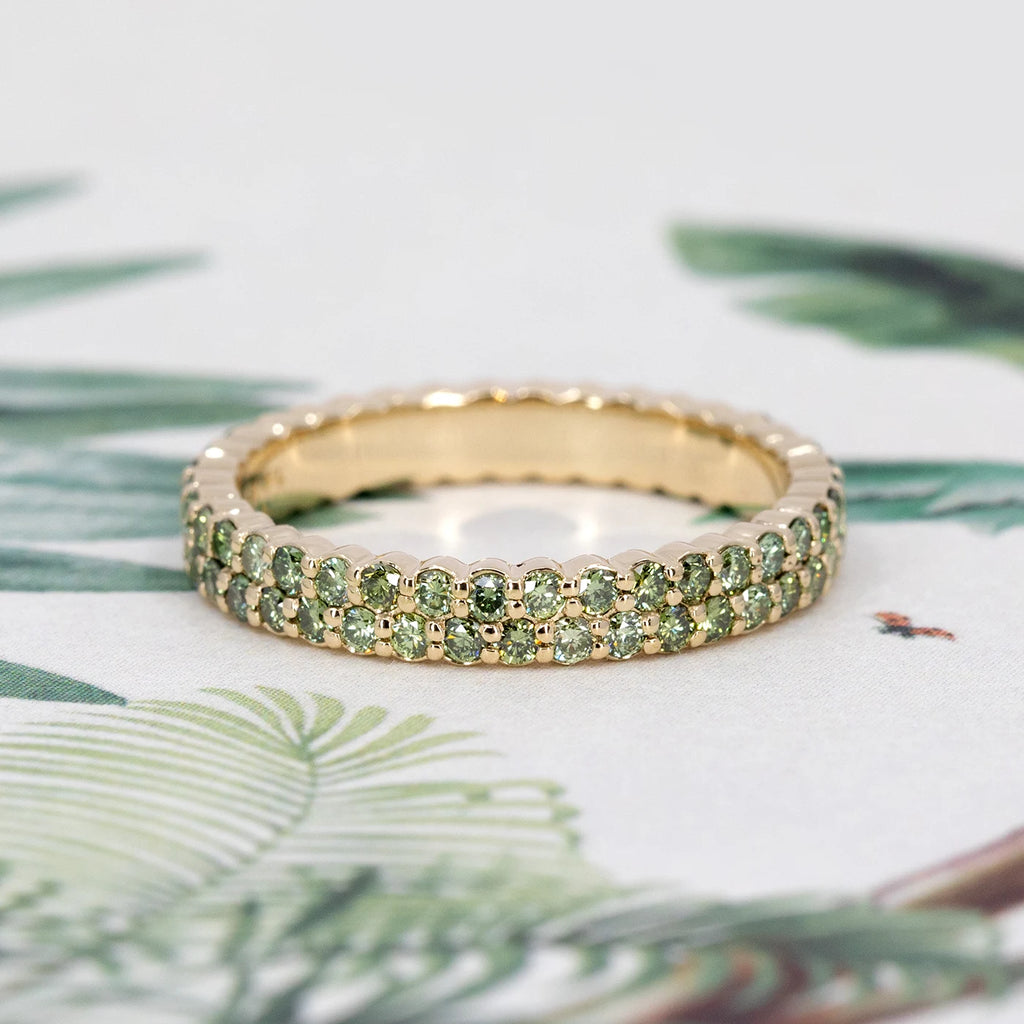 Green diamond eternity ring with double rows of pavé-set stones in 14k gold, showcased against a soft tropical-inspired background. Independent Canadian jewelry from Ruby Mardi, Montreal.