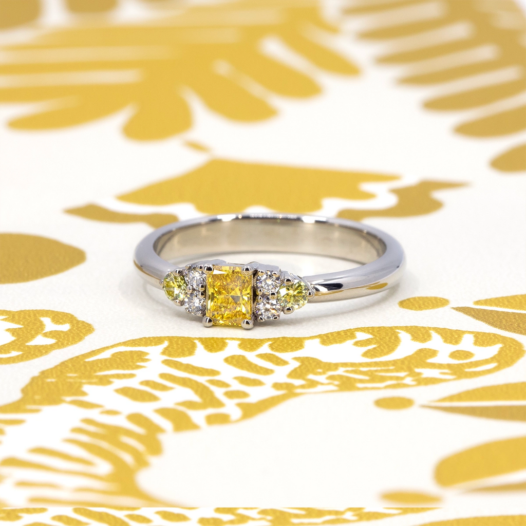 Radiant yellow lab-grown diamond ring with canary yellow and white diamond accents in 10k white gold, displayed on a yellow and white patterned background – Ruby Mardi Montreal.