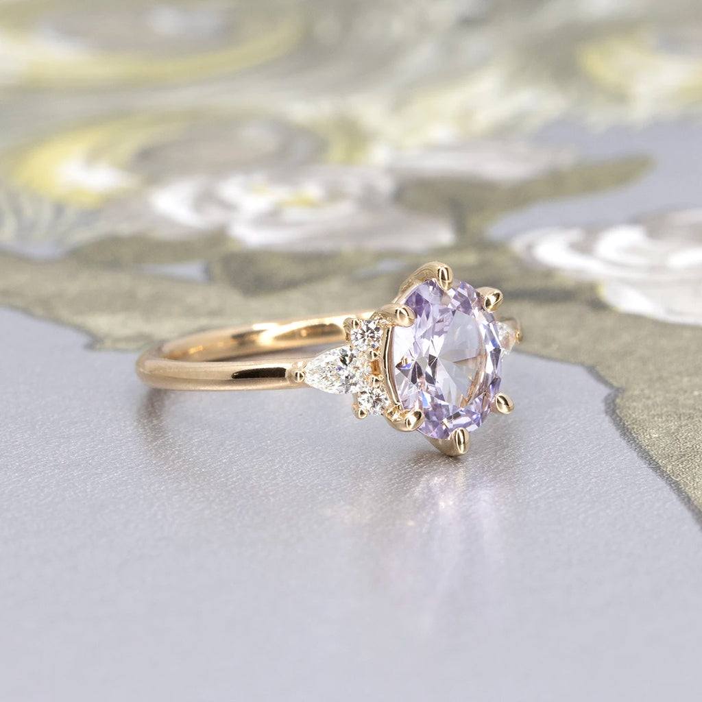Lilac sapphire engagement ring with pear-cut and round diamond accents in 14k yellow gold, photographed on a floral patterned background. Handmade in Montreal by Maksym Jewelry, available at Ruby Mardi.