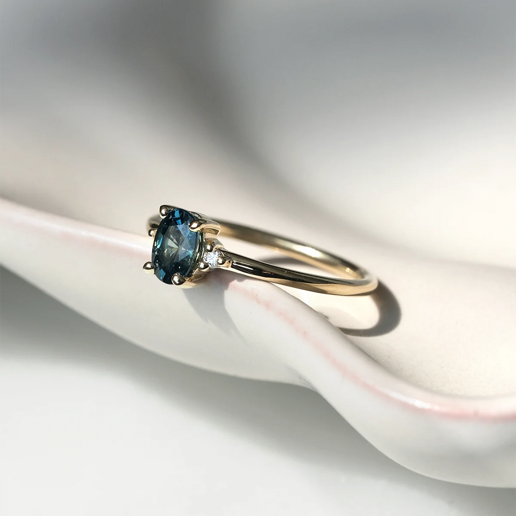 Yellow gold engagement ring with oval teal sapphire and tiny side diamonds, handmade by Maksym Jewelry in Montreal. Photo shows the ring in natural light on a porcelain shell dish. Available at Ruby Mardi, the coolest jewelry store in Canada.