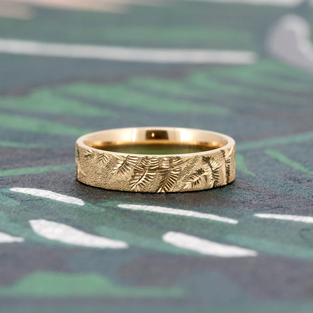 14k yellow gold wedding band hand-engraved with delicate fern motifs by Rebel & Rogue, presented on a botanical green background. The stippled texture creates a rich contrast with the foliage engraving. Shot for Ruby Mardi in Montreal, Canada, showcasing independent designer fine jewelry.