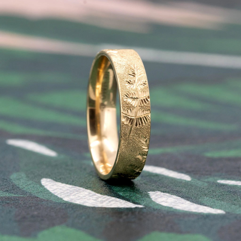 Hand-engraved fern motif gold band in 14k yellow gold by Rebel & Rogue, photographed on a textured green patterned surface. Made for Ruby Mardi in Montreal, Canada, featuring independent designer craftsmanship.