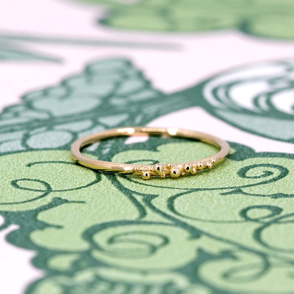The Areia ring in 14k yellow gold by Meg Lizabet, shown against a green floral background. The band features a delicate row of organic gold granules. Available at Ruby Mardi in Montreal.