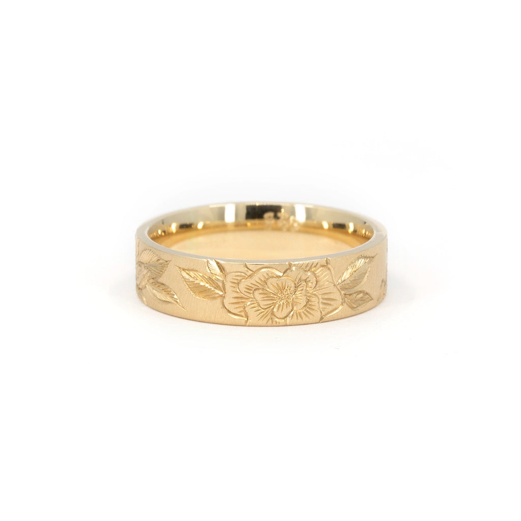 Detailed close-up of a handcrafted gold band with deeply carved floral engravings and textured background. The ring rests on a clean white surface, showcasing the precision of Rebel & Rogue’s hand engraving, available at Ruby Mardi in Montreal.