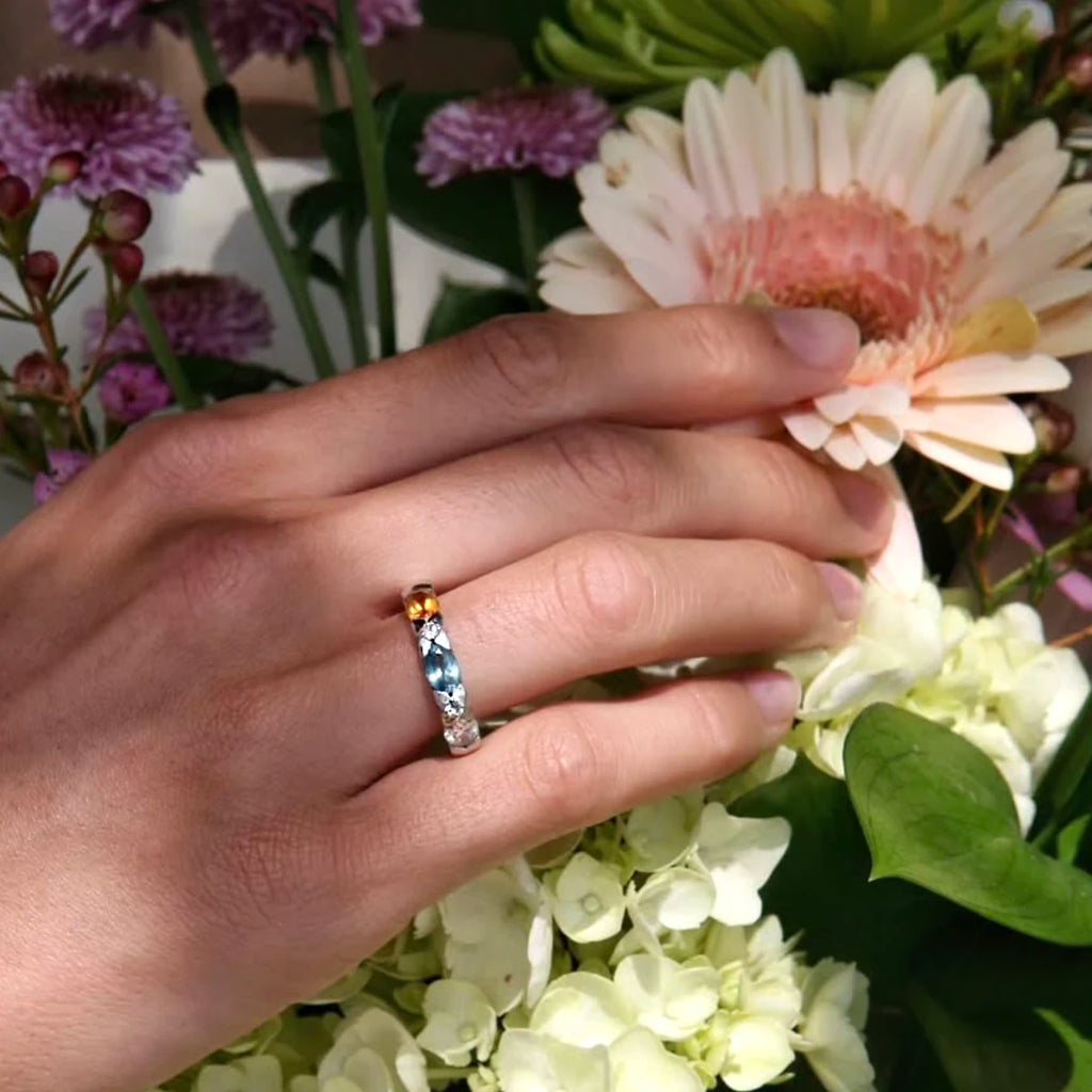Hand wearing the Myosotis ring by Fleurs Sauvages QC, featuring citrine, London blue topaz, lab-grown diamond, and colorless sapphires set in 18k white gold, photographed among fresh summer flowers at Ruby Mardi in Montreal.