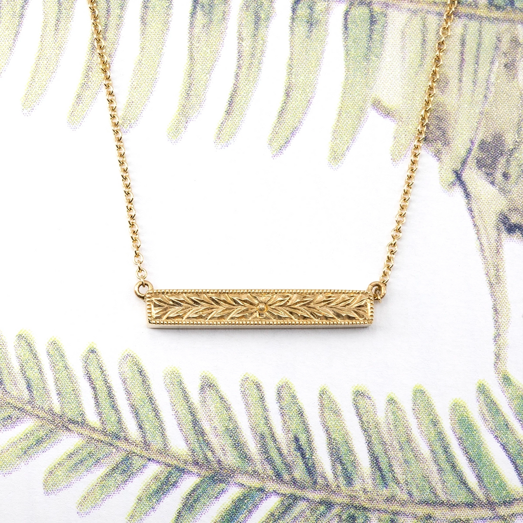 Ruby Mardi fine jewelry – 14k yellow gold bar necklace with intricate floral and leaf engraving. Elegant handcrafted pendant with vintage-inspired details, displayed on a botanical background.