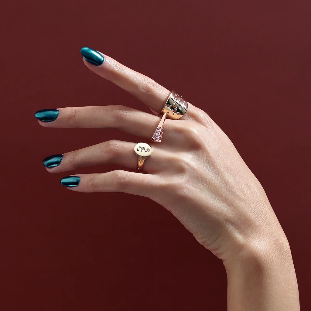 A hand with dark green blue metallic nails is photographed in studio on a red wine color background. The hand wears 3 designer rings. 2 of them are yellow gold with diamonds, and the one that's on the middle finger is in rose gold with a pink sapphire pavé. All these fine jewelry pieces are available at Ruby Mardi, the best independent jewelry store in Canada.
