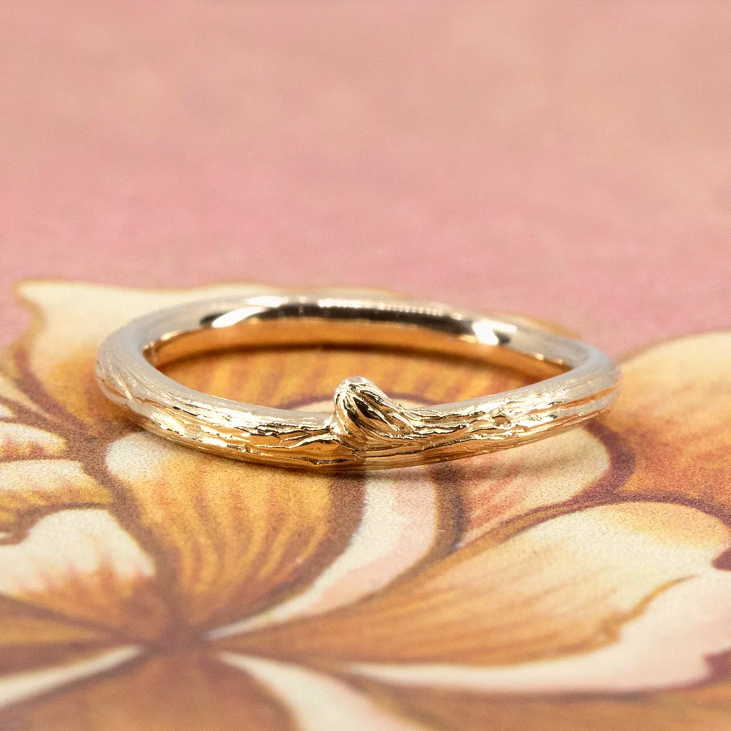 Gold wood-textured ring by Meg Lizabet, photographed on a soft floral illustration. The organic band in solid yellow gold features a sculpted knot and bark-like texture, evoking a delicate tree branch. Handmade in Montreal and available at Ruby Mardi.