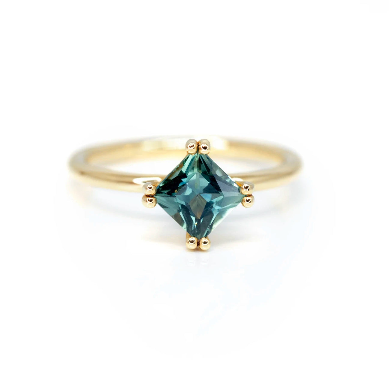 A princess cut sapphire set in yellow gold with elegant double prongs is seen on a white background. This designer engagement ring was handmade by Sheena Jewelry in Montreal. It’s the perfect ring for stacking. Find more bridal rings at the best jewelry store in Canada, Ruby Mardi.