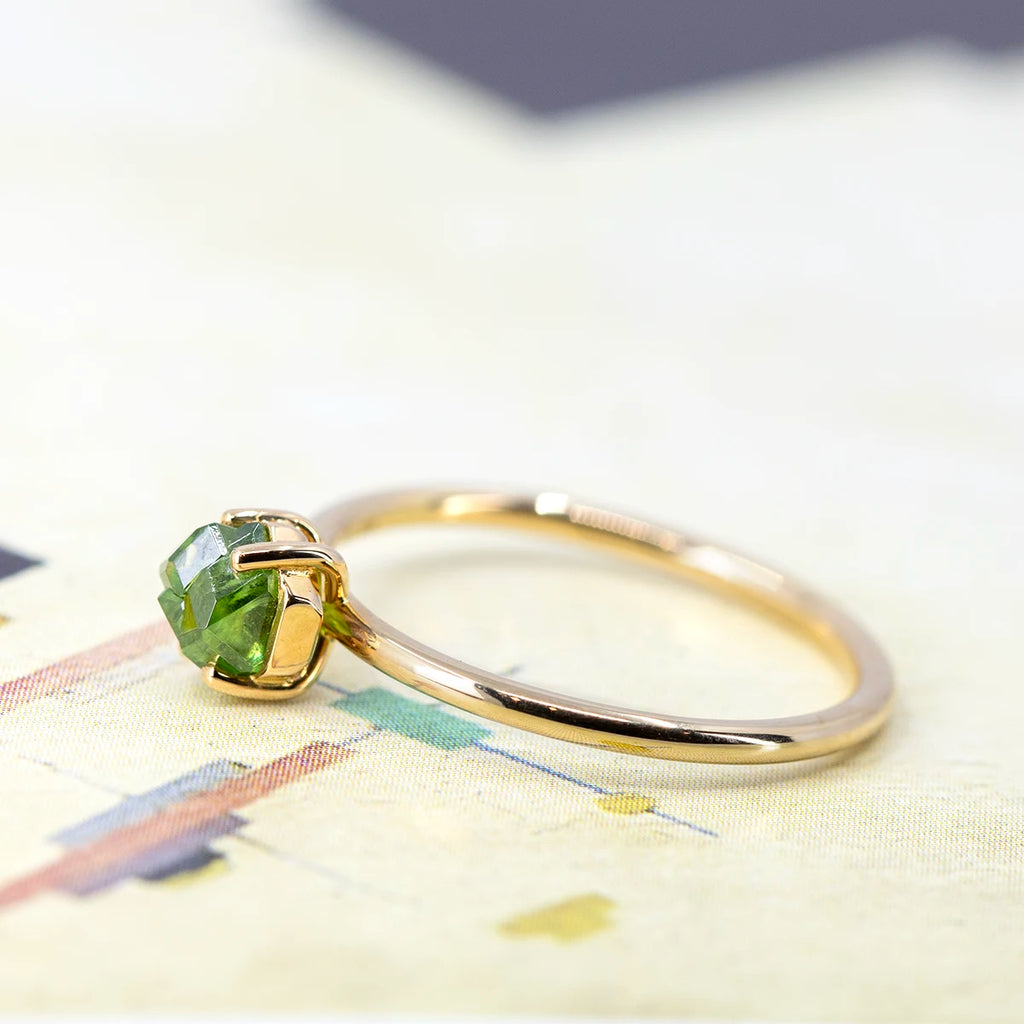 A bridal ring in 18k yellow gold featuring a rough green gemstone (demantoid garnet from Quebec) handmade in Montreal by independent jeweller François Charest in collaboration with boutique Ruby Mardi, a fine jewelry store in Montreal. The ring is seen photographed on a cubic art background.