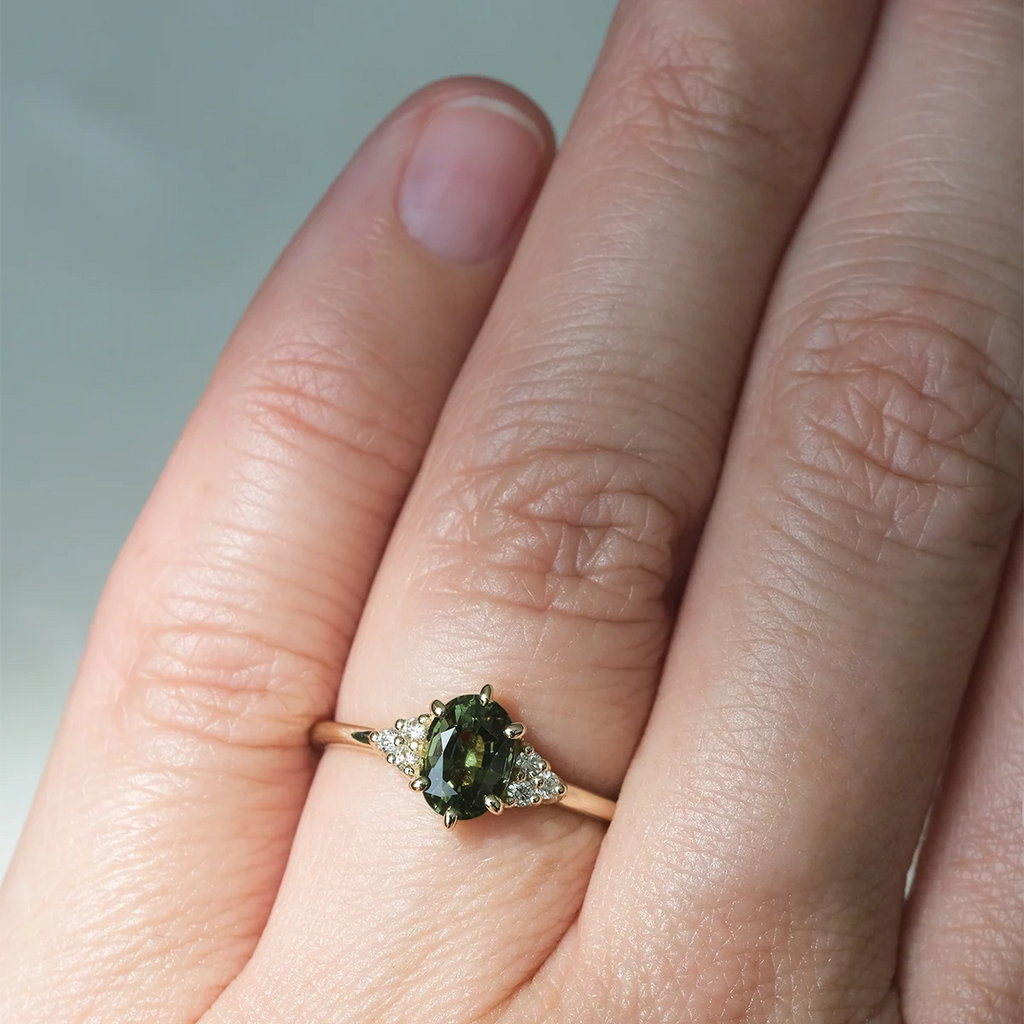 Green sapphire engagement ring on hand, crafted in yellow gold with side diamonds. Maksym Jewelry design, available at Ruby Mardi, Montreal.