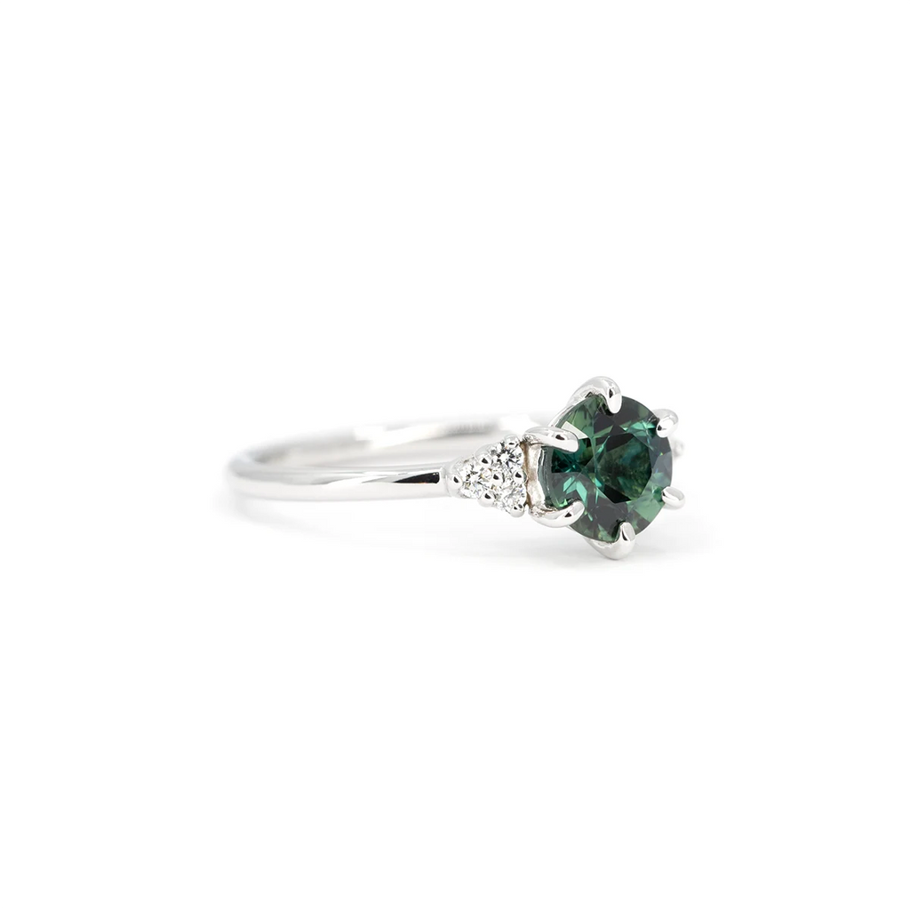 Close-up of a teal sapphire engagement ring with diamond side accents in white gold, photographed on a white background. Designer ring by Maksym Jewelry, available at Ruby Mardi, Montreal.