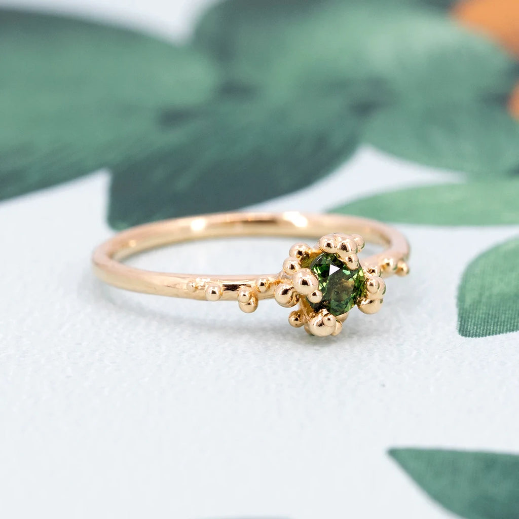 A unique green sapphire solitaire ring with organic gold bead details, displayed on a leafy background. Designed by Meg Lizabet and available at Ruby Mardi in Montreal, this handcrafted piece exemplifies artisanal elegance and creativity.