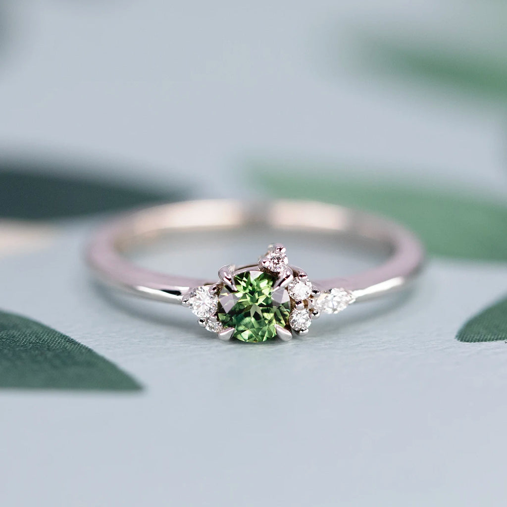 White gold engagement ring featuring a round green sapphire accented by delicate clusters of diamonds, displayed against a soft botanical background. Available at Ruby Mardi in Montreal.