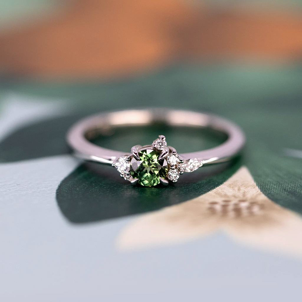 Delicate white gold engagement ring with a round green sapphire and small clustered diamonds, displayed on a soft botanical illustration. Available at Ruby Mardi in Montreal.