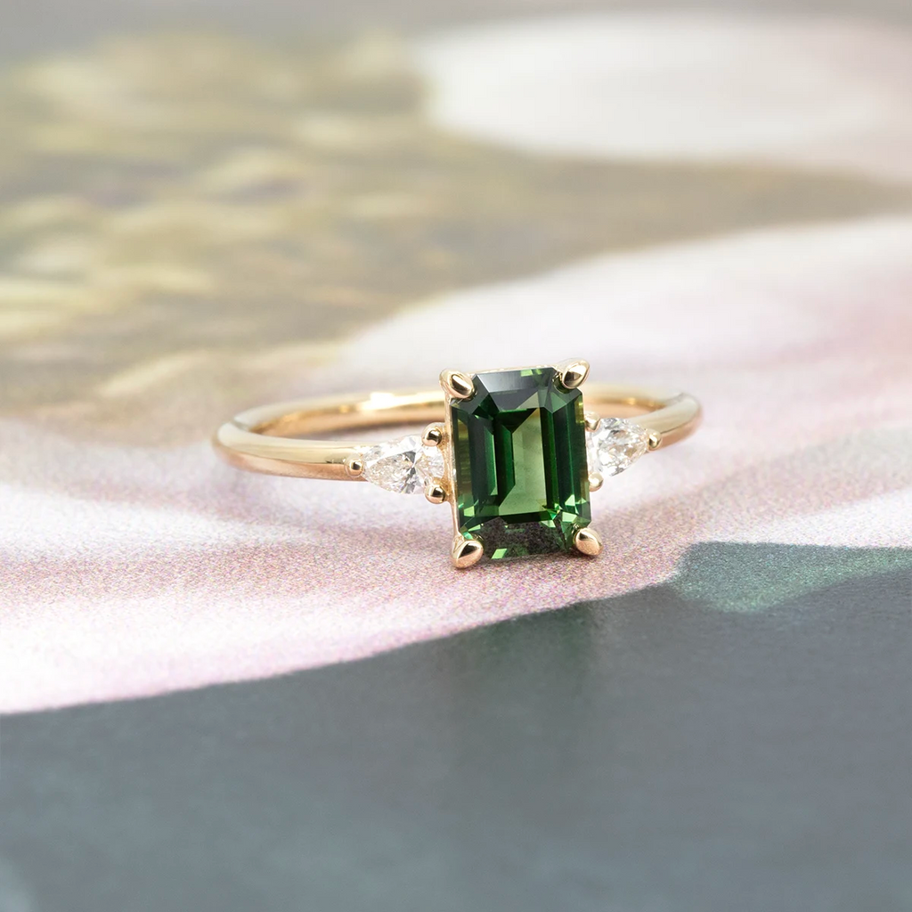 Three-quarter view of a yellow gold engagement ring featuring an emerald-cut green sapphire and pear-shaped diamond accents. Set on a soft, floral surface. Crafted by Maksym Jewelry in Montreal, available at Ruby Mardi.