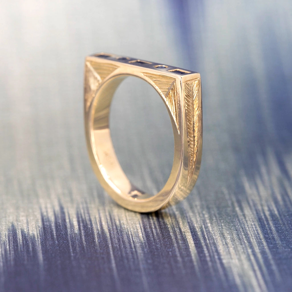 Hand-engraved 14k yellow gold ring with deep blue baguette and round sapphires, Rebel & Rogue Jewelry, photographed upright on a textured blue surface. Available at Ruby Mardi, best jewelry store in Canada.