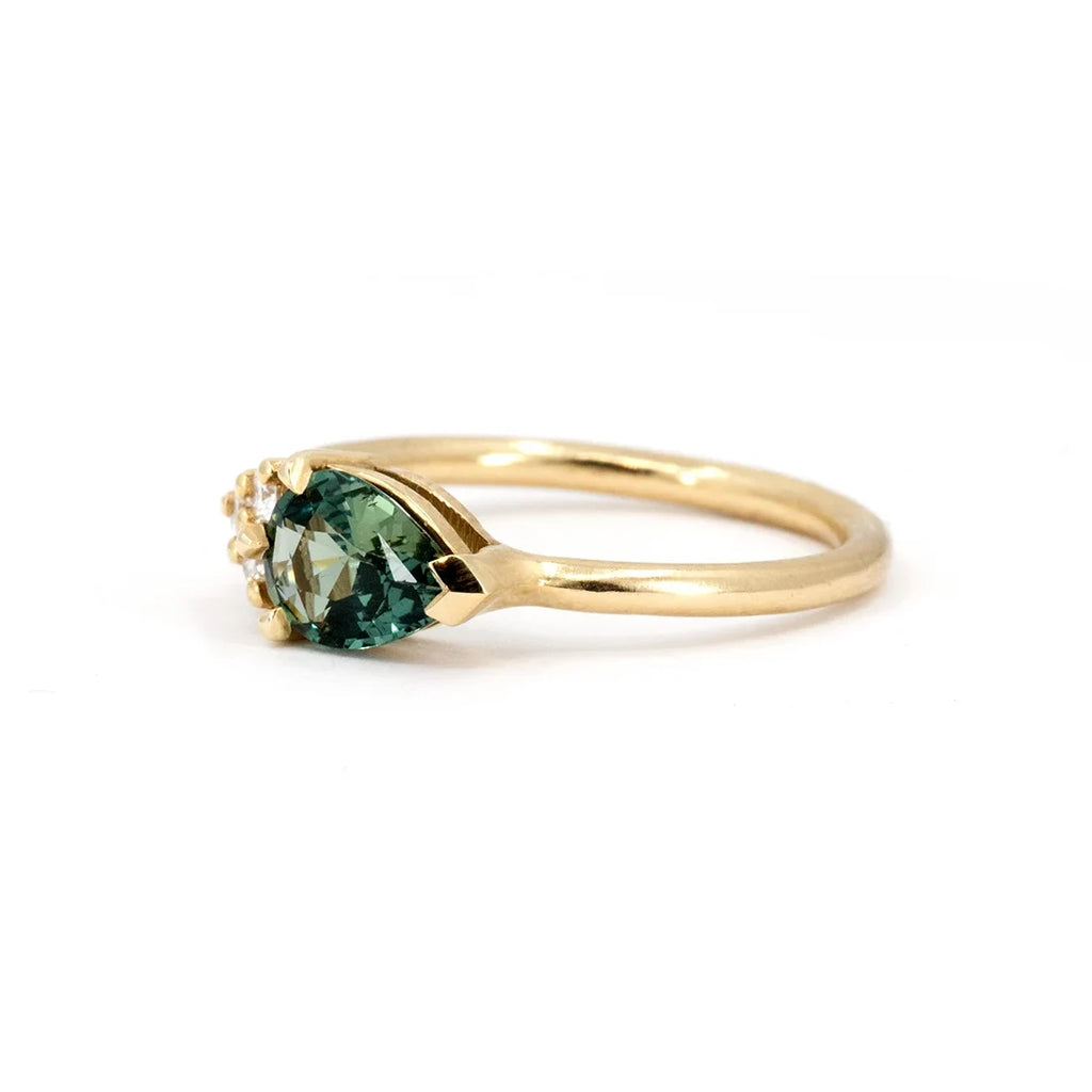 Handcrafted engagement ring featuring a pear-cut teal sapphire and diamond accents, set in polished yellow gold. Designed by Sheena Purcell for Ruby Mardi, this elegant piece showcases fine craftsmanship and modern elegance. Made in Montreal, Canada.