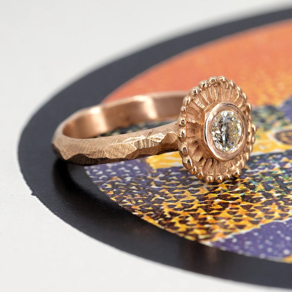 A side view on a colored background of a fairly solid rose gold jewel with a raw and “organic” style. The band itself is wide and textured. In the center, a natural round white diamond in a bezel setting, around which the rose gold forms a sunburst “halo”. This jewel is available only from Ruby Mardi, a luxury jewelry store that, like Gem Breakfast, sells the work of independent jewellery designers. 