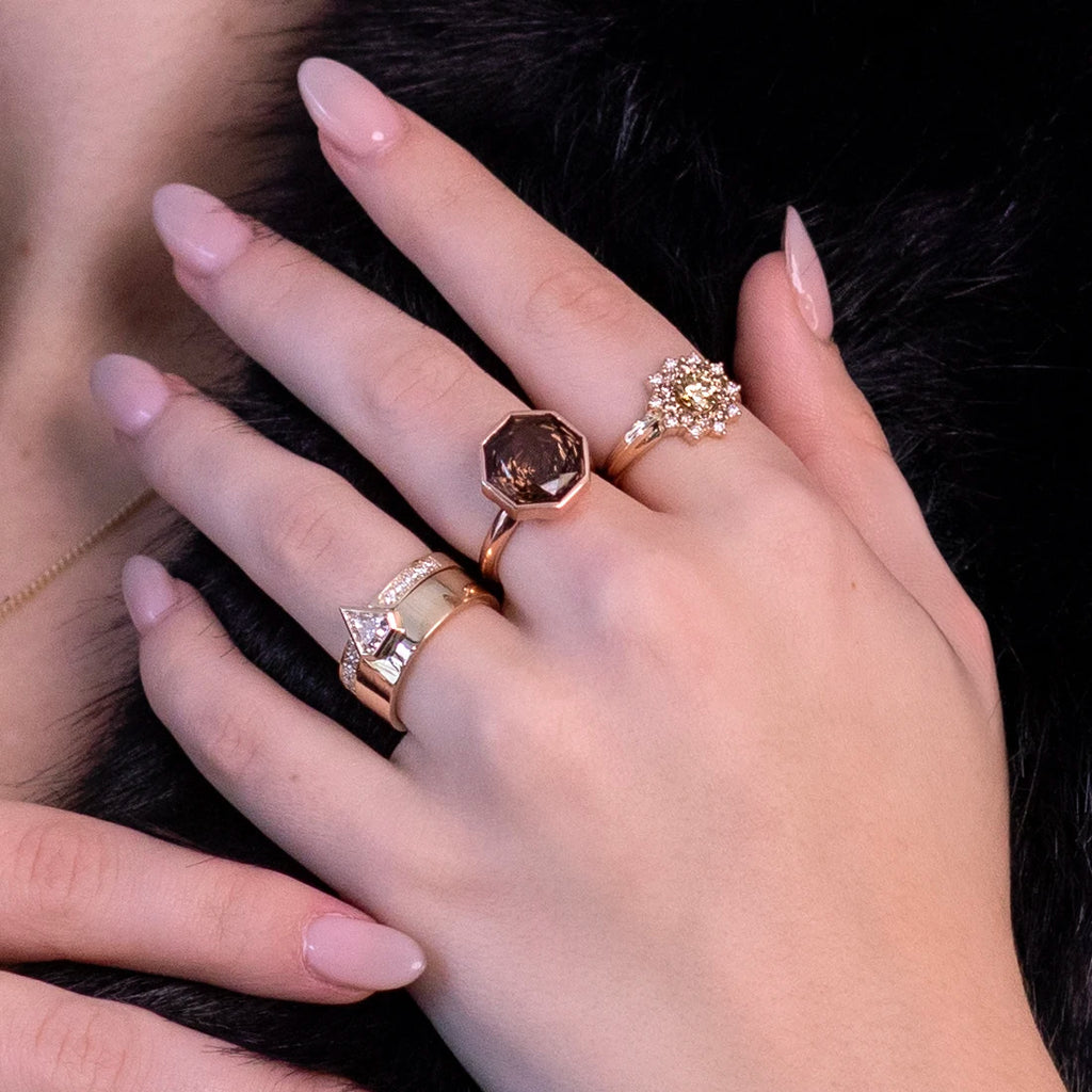 A close-up of a hand adorned with elegant rings, including an octagon smoky quartz cocktail ring by Bena Jewelry in a rose gold bezel, a modern gold wedding set with geometric details, and a dazzling yellow diamond cluster ring. These unique, handcrafted designs are available exclusively at Ruby Mardi, a boutique specializing in artisan jewelry.






