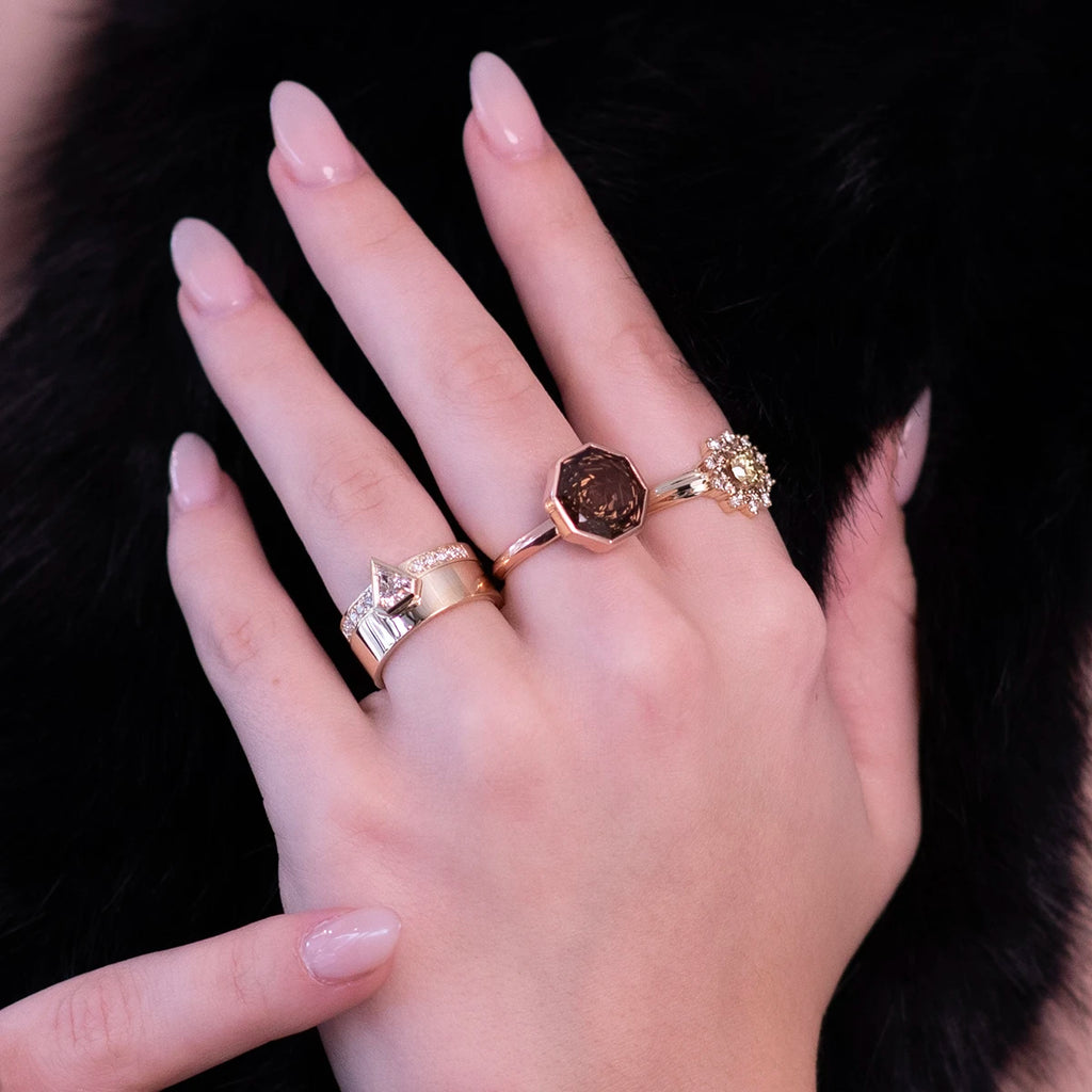 A close-up of a hand adorned with three luxurious rings: a bold yellow gold ring with a pentagonal diamond, an octagonal ring with a brown gemstone, and a gold ring featuring a floral cluster of diamonds. The nails are polished in a nude shade, and the background of soft black fur enhances the jewelry's elegance and sophistication.