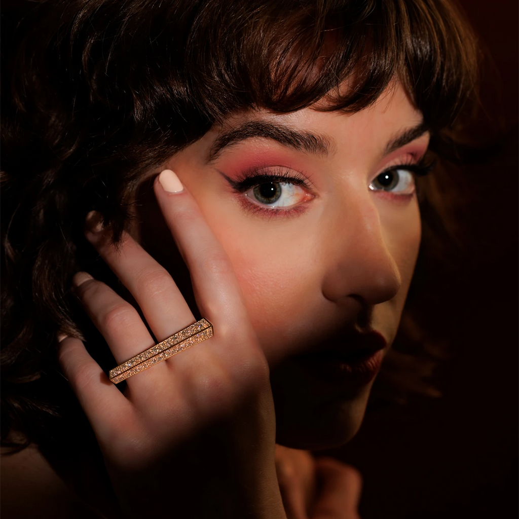 Close-up photograph of a woman's face with brown hair and mouth in shadow. The eyes (made up in red with a pronounced line of eyeliner) are lit up and look out at the camera. Near her eyes, she brings her hand to her face, on which she wears a luxurious piece of high-end jewelry: a 14-kt gold ring set with 2.37 carats of diamonds. It's a long, generous gold ring that can be worn on two fingers. A modern, fashionable and unisex piece of jewelry, created by independent Canadian designer Bena Jewelry.