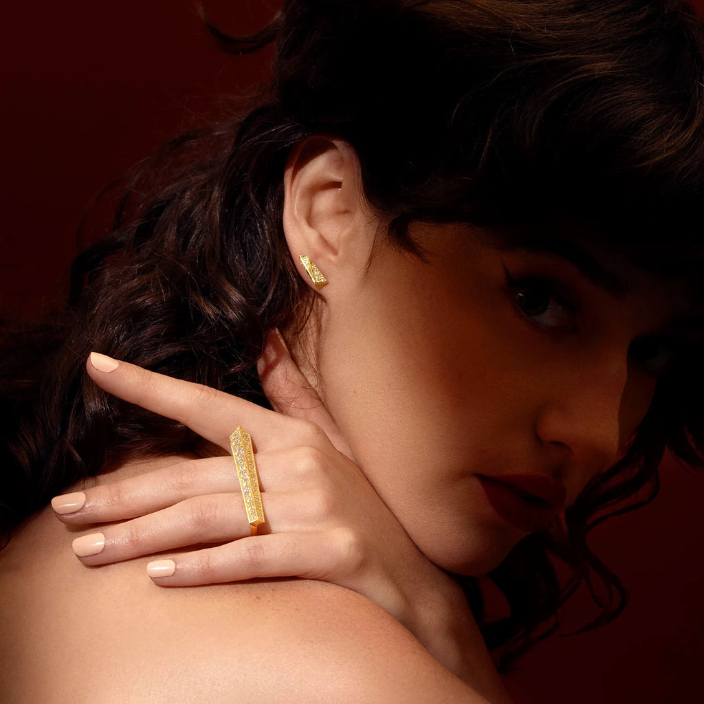 Fashion photography of a young woman with curly brown hair whose face is in shadow. She looks at the camera and raises her hand over her shoulder: on her hand, illuminated by the light, she wears a unique high end ring that looks like an objet d'art. It's a large yellow gold baguette set with several diamonds. She is also wearing a matching diamond earring. The jewels have a modern, geometric look, created by independent Canadian designer Bena Jewelry.