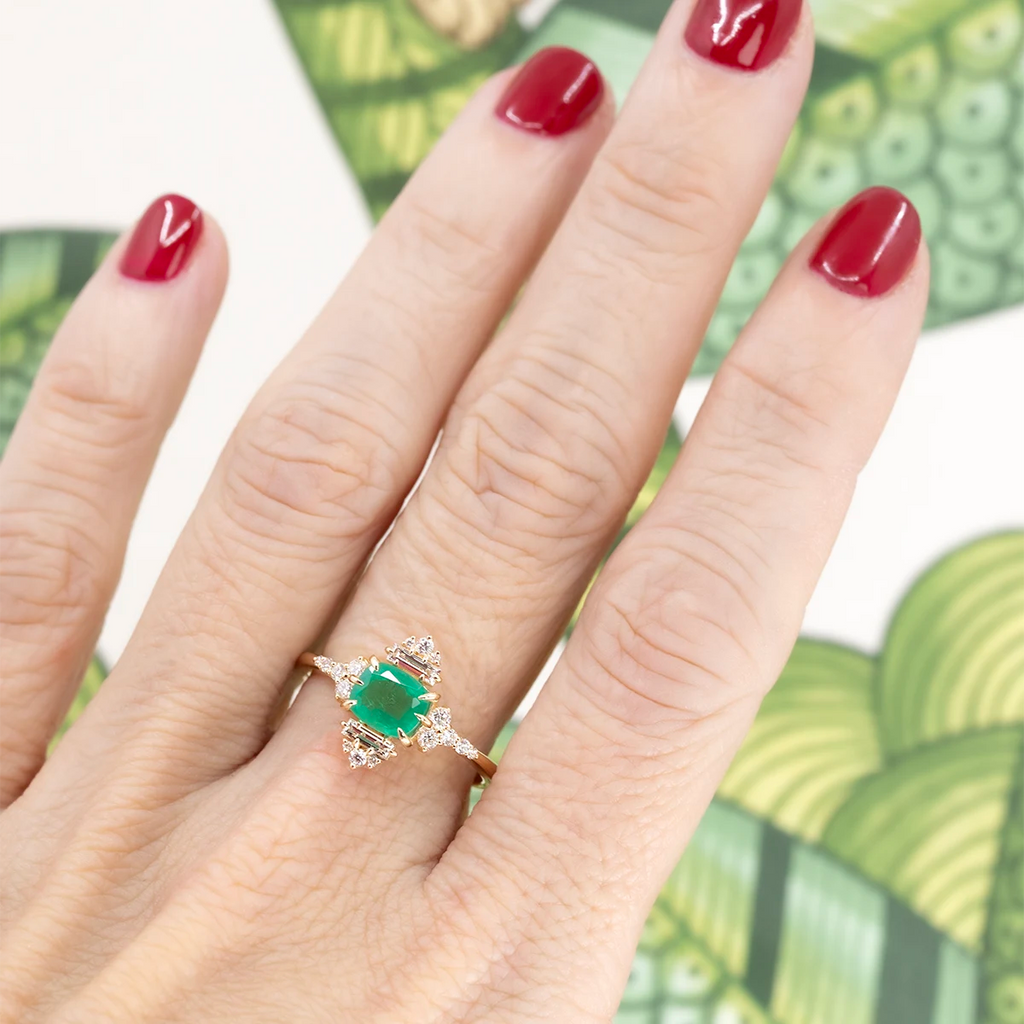 A high end piece of jewelry worn by a hand with red nails. The ring features a central genuine emerald with accent diamonds. This designer engagement ring or right hand ring is available at Ruby Mardi, the best jewelry store in Montreal.