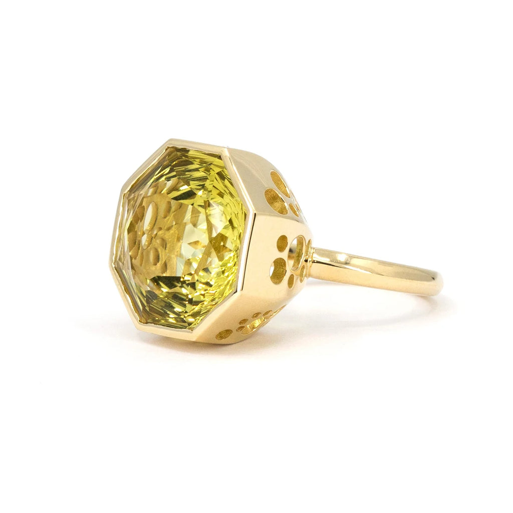 A large ring seen from the side, photographed against a white background. The large yellow gold basket is pierced by circular holes that let the light into the yellow-green gemstone. The gemstone is so large that the ring band is barely visible behind it. It is set in 14-carat yellow gold. This is a unique creation by Canadian jewelry designer Bena Jewelry. 