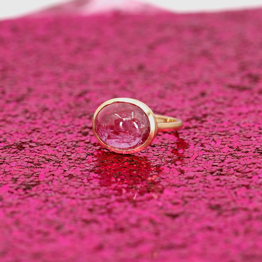 A big pink gemstone ring is photographed on a background made of bright pink glitters. Pink on pink with a gold frame around the naturally included gemstone. Find this gem of a jewelry piece at Ruby Mardi in Montreal.