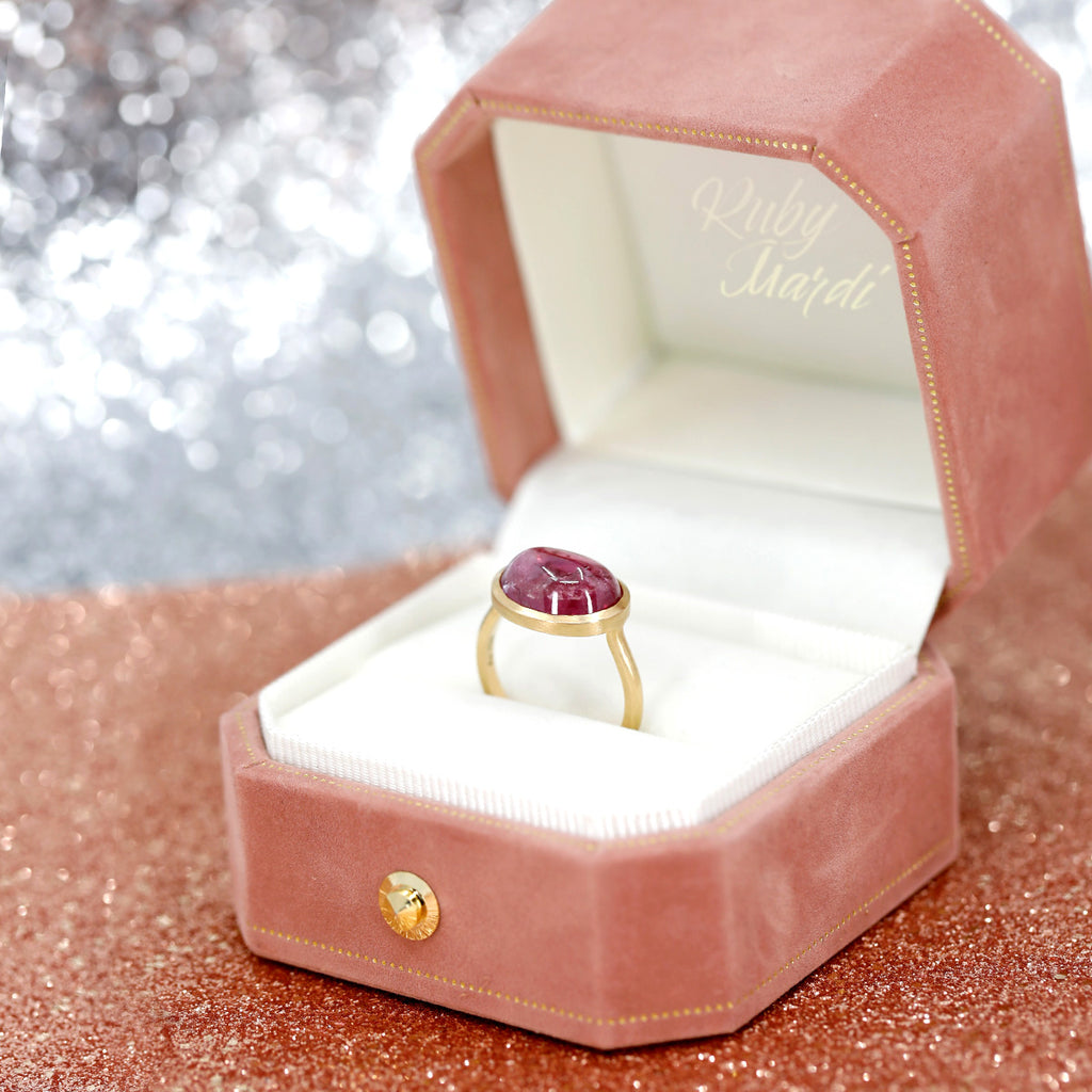 A big ruby cabochon ring set in 18k yellow gold is seen photographed on a pink velvet box with a Ruby Mardi tag, the name of the jewelry store that sells this one-of-a-kind right hand ring. The jewelry box is photographed on a pink sequins background. 