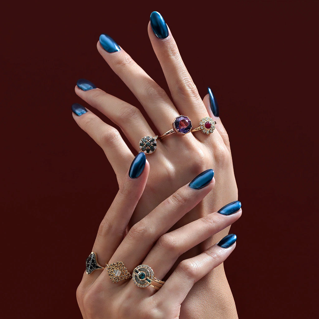 Two lady hands with metallic blue nails are photographed on a red background and wear one-of-a-kind fine jewellery pieces. We can see six statement gemstone rings or black diamond rings, with all different styles. All these high end rings can be found at Ruby Mardi, an independant jewelry store in Canada selling the work of independent jewellers, as Gem Breakfast does.
