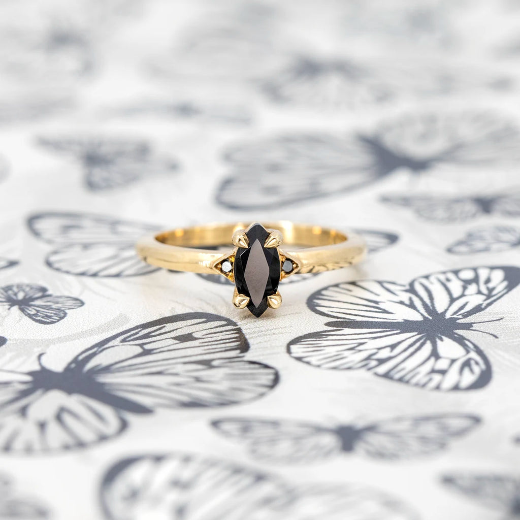 Marquise black gemstone ring in yellow gold with unique claw prongs, designed by Canadian jeweller Liane Vaz, photographed on a butterfly-patterned background.