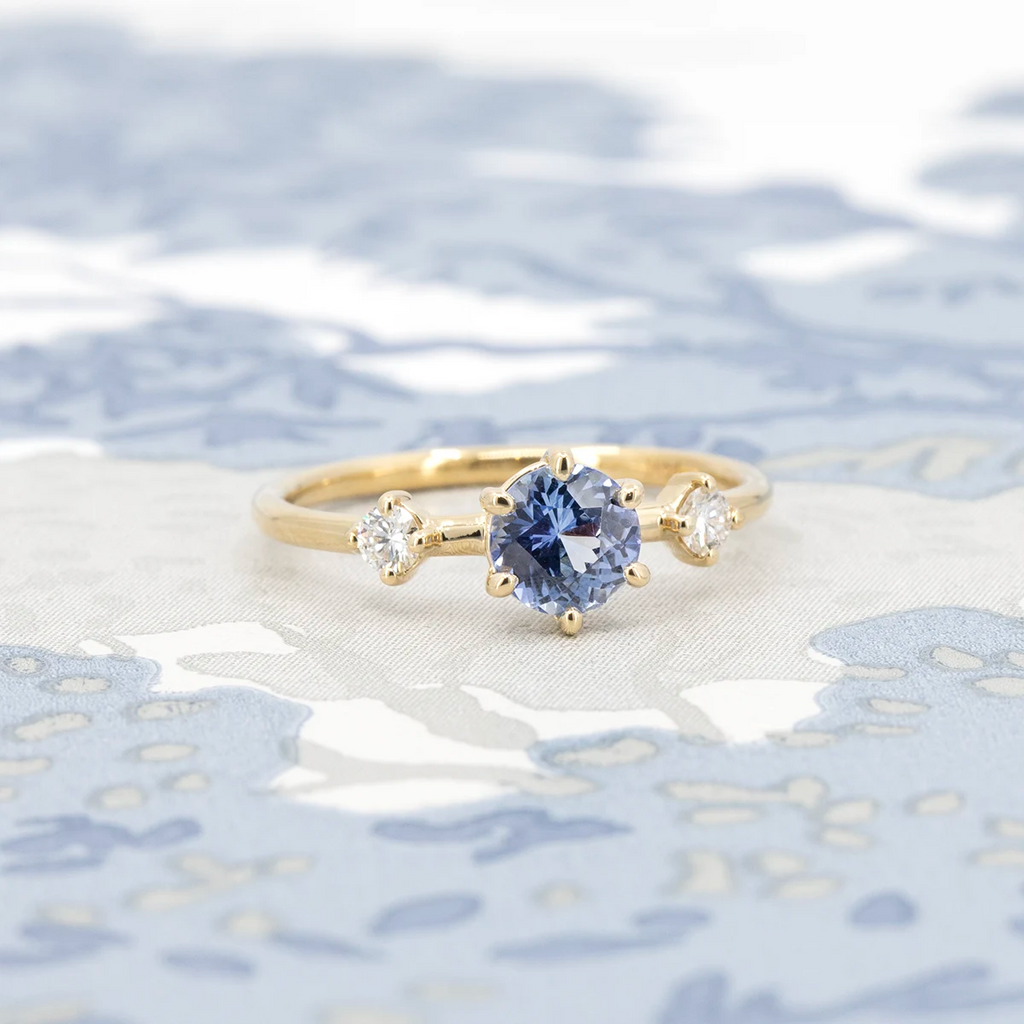 Isabella light blue sapphire and diamond engagement ring in yellow gold, handcrafted by Maksym Jewelry in Montreal. Displayed on a blue and white floral background. Available at Ruby Mardi.