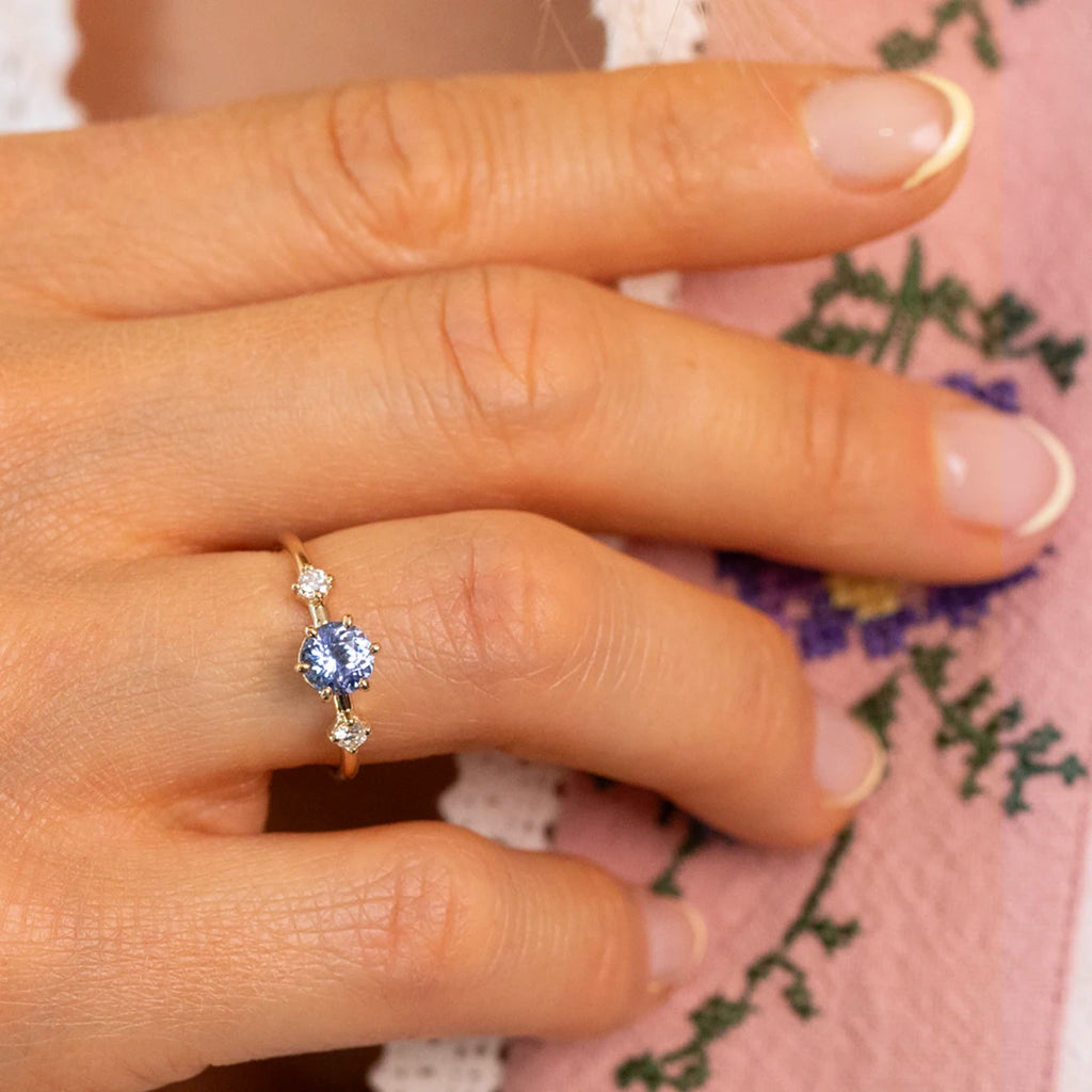 Close-up of hand wearing Isabella ring with light blue sapphire center and diamond accents on yellow gold band, alternative engagement ring by Maksym for Ruby Mardi.