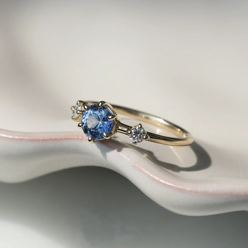 Isabella engagement ring with light blue sapphire and white diamonds in yellow gold, handmade in Montreal by Maksym Jewelry. Photographed on a sculpted ceramic dish. Available at Ruby Mardi.