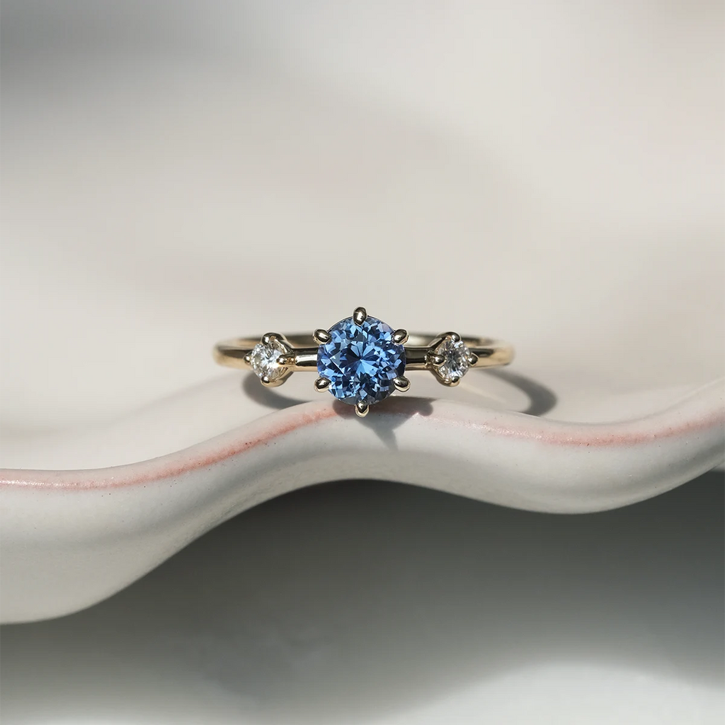 Light blue sapphire and diamond engagement ring in yellow gold, handmade by Maksym Jewelry in Montreal. Shown on a ceramic dish, available at Ruby Mardi.