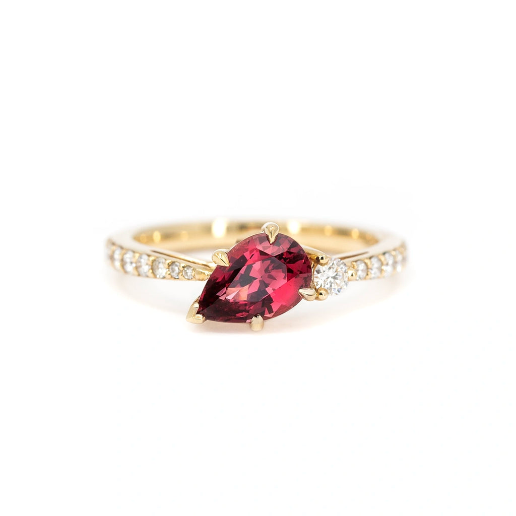 A gold ring handcrafted by Justine Quintal, featuring a pear-shaped red spinel gemstone set at an angle, accented by a single round white diamond and a band adorned with small pavé-set diamonds, displayed on a white background.