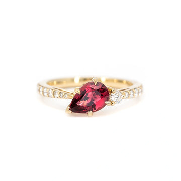 A gold ring handcrafted by Justine Quintal, featuring a pear-shaped red spinel gemstone set at an angle, accented by a single round white diamond and a band adorned with small pavé-set diamonds, displayed on a white background.