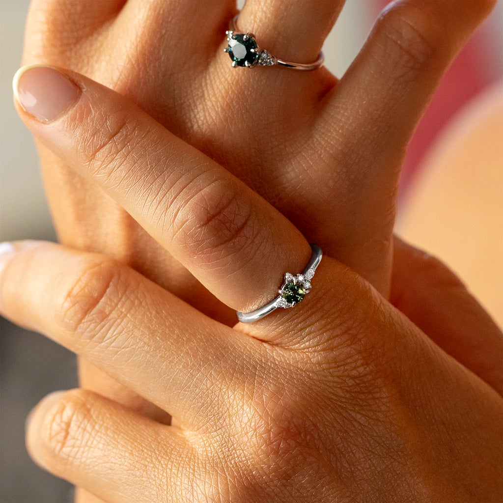 White gold engagement ring with a round dark green sapphire and small diamond accents, part of the Little Folie collection by Justine Quintal, shown worn on the hand.