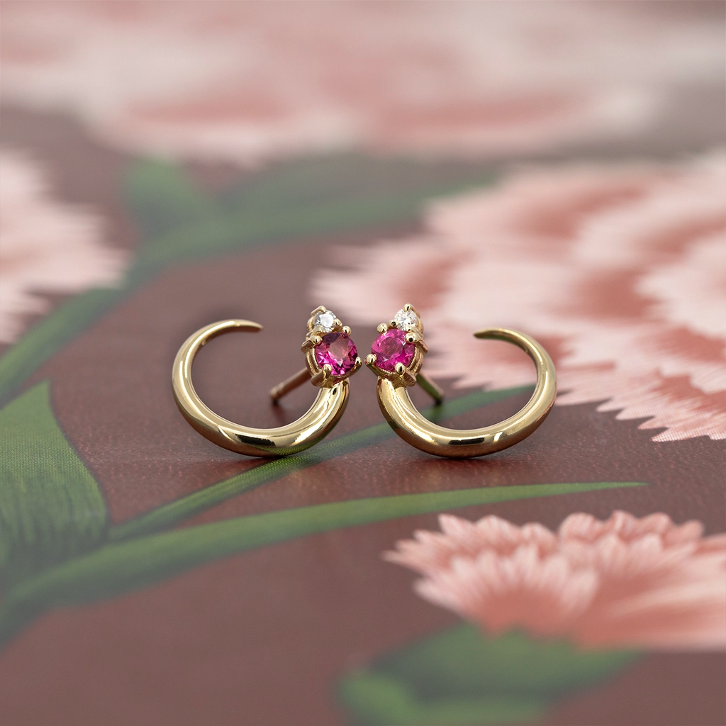 Pair of gold crescent earrings with vivid pink tourmaline and small white diamond accents, designed by Justine Quintal, contemporary fine jewelry, photographed on a floral background, Montreal, Canada.