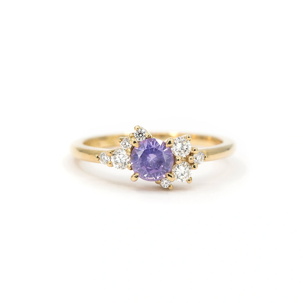 A gold ring featuring a round purple sapphire at the center, accented with asymmetrically arranged small round white diamonds on each side, displayed on a white background.