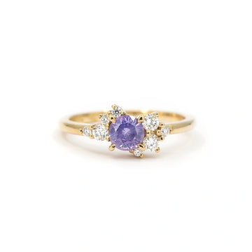A gold ring featuring a round purple sapphire at the center, accented with asymmetrically arranged small round white diamonds on each side, displayed on a white background.