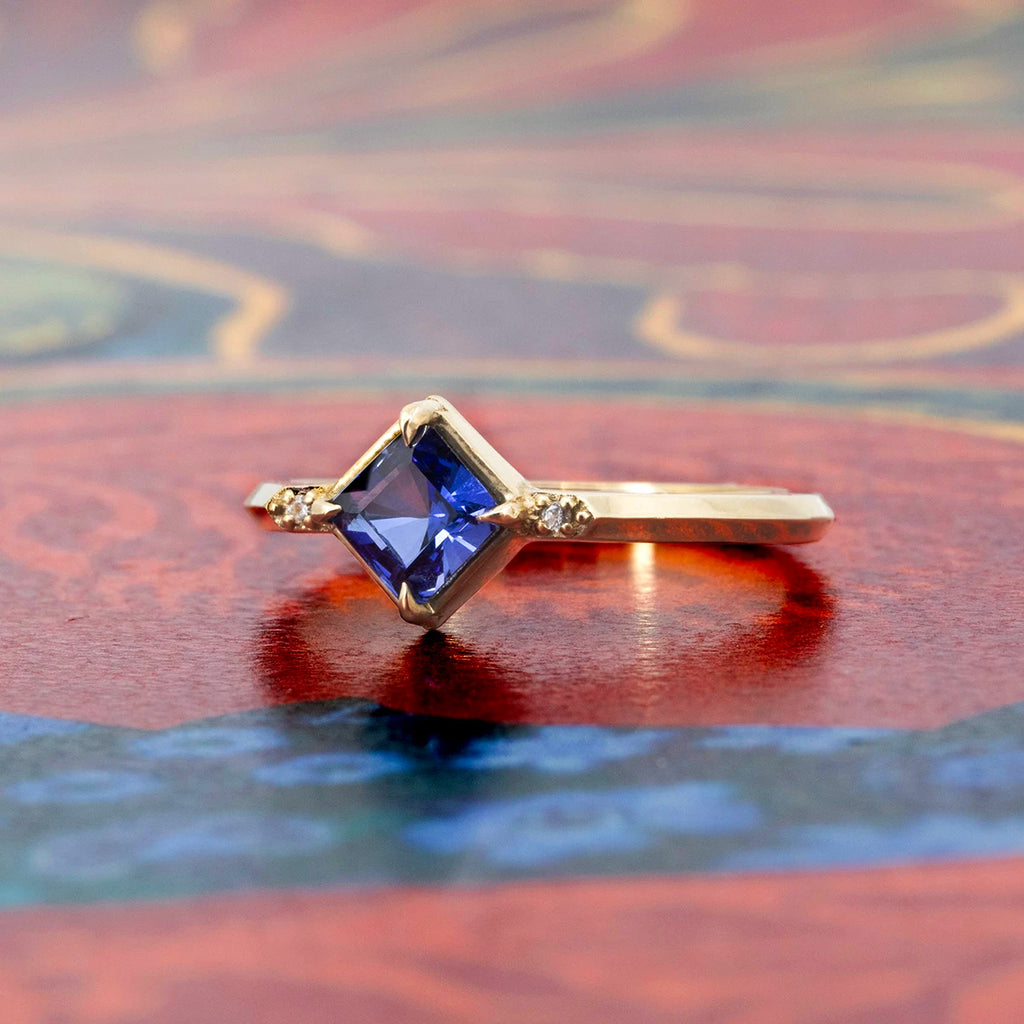 Square blue sapphire ring in yellow gold with diamond accents, minimalist engagement ring by Liane Vaz for Ruby Mardi, displayed on colorful background.