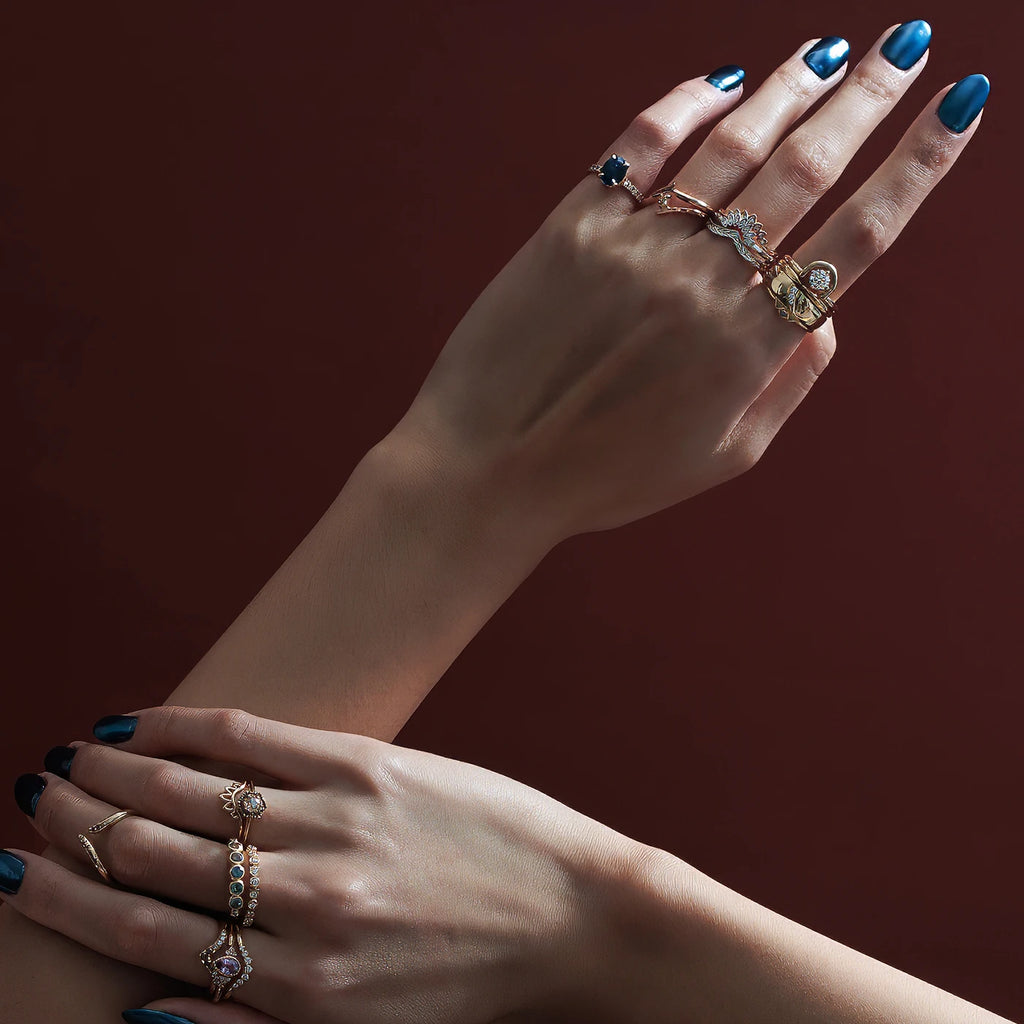 A fashion shoot of two hands with a blue metallic manucure wearing multiple designer rings. Diamond wedding bands and sapphire engagement rings are seen.