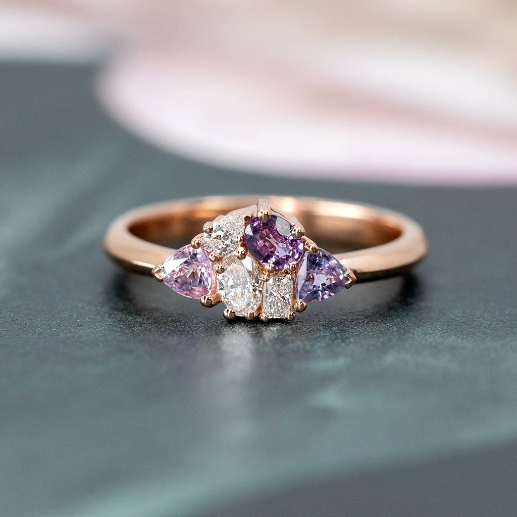 A close-up view of a 14k rose gold cluster ring with three light purple sapphires and three lab-grown diamonds, set against a soft pastel background with muted pink and forest green floral motifs. The ring's romantic, vintage-inspired design is beautifully highlighted by the delicate, nature-inspired backdrop.