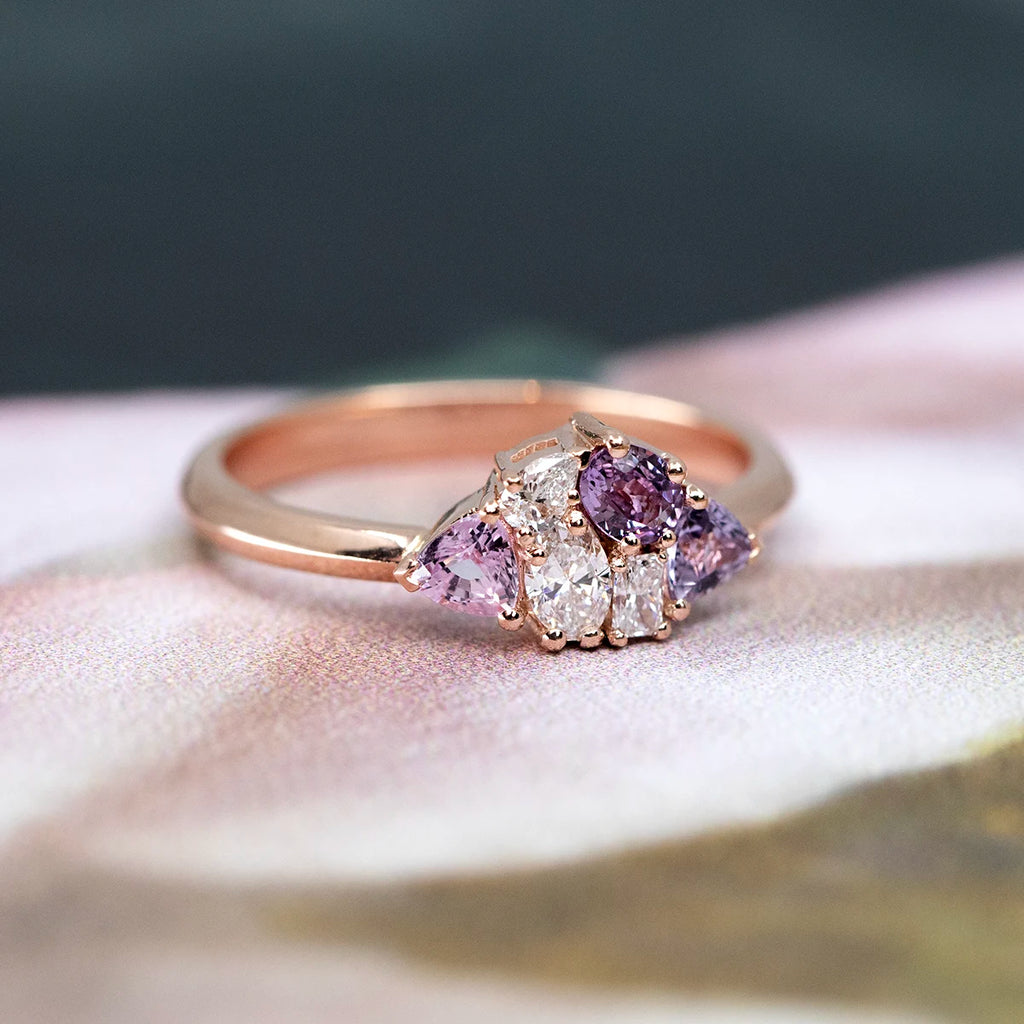 A 14k rose gold cluster ring with three light purple sapphires and three lab-grown diamonds, photographed from the side on a soft, floral-inspired background with pastel pink and forest green hues. The romantic setting enhances the ring’s delicate and vintage-inspired charm.