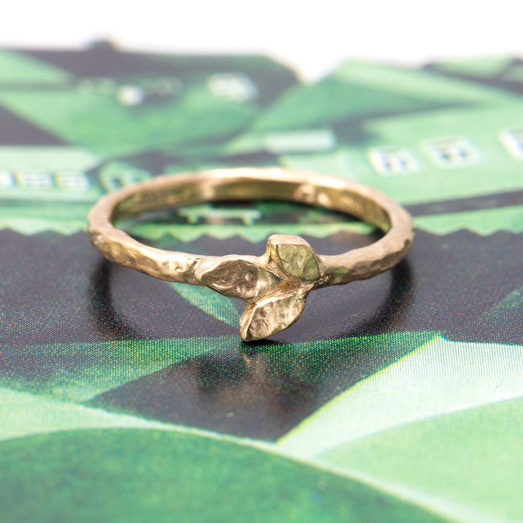 Original wedding band in yellow gold with a hammered textured is photographed in close up on a green abstract background. The ring features three gold leaves on the center of the band. It’s a gold band with a rough texture. It was handcrafted and is available at fine jewellery store Ruby Mardi in Montreal.